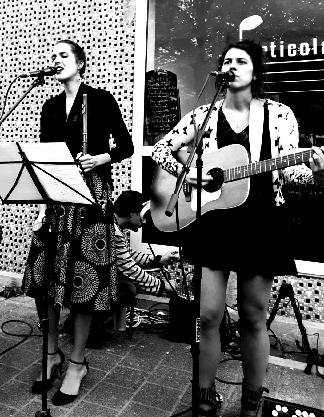 Alissia and Morgane from Lyonesse on an outdoor stage, Alissia holding her flute and Morgane her guitar, both singing.